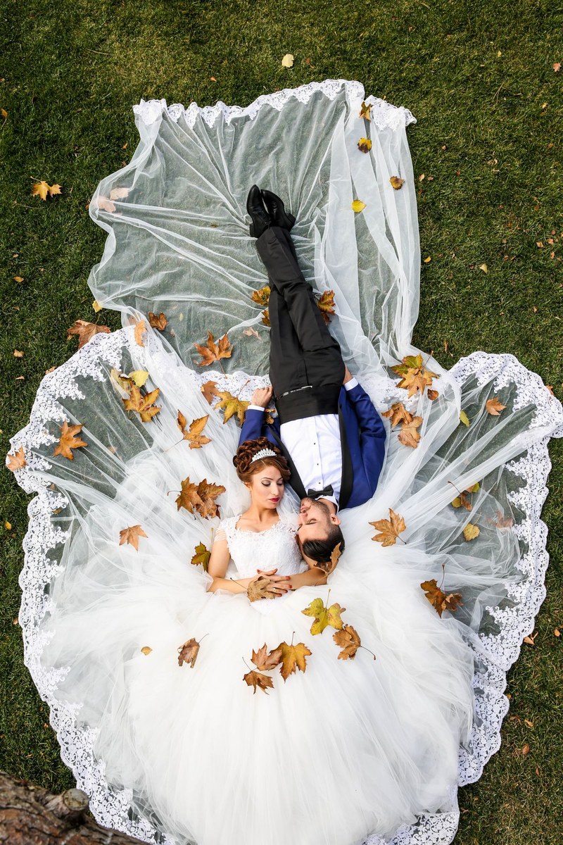 Bride and groom lying on grass with her lace gown and veil spread out among autumn leaves